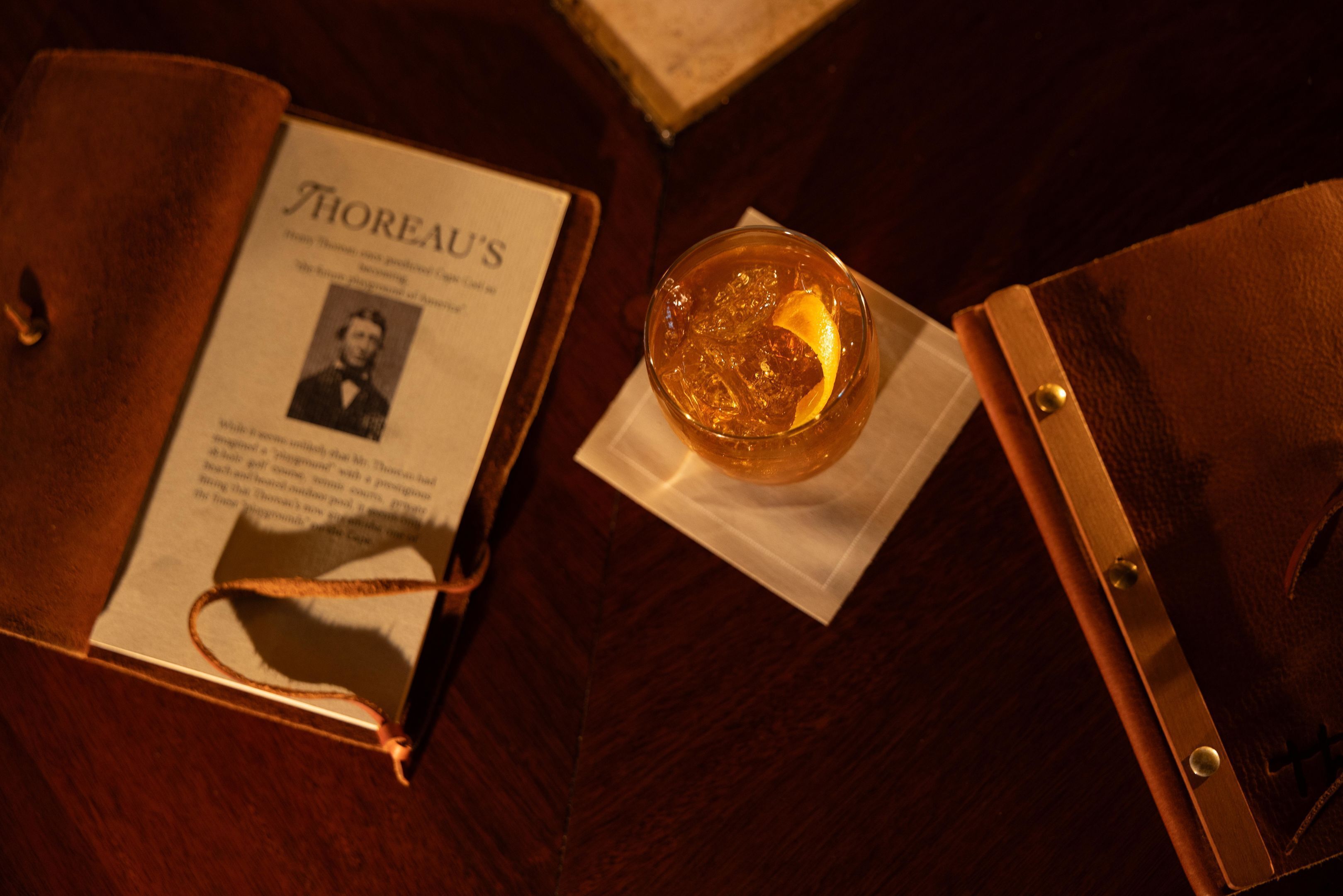 A book titled 'Thoreau's' with a photograph, a glass of amber liquid with ice and a lemon slice, and a wooden folder on a wooden surface.