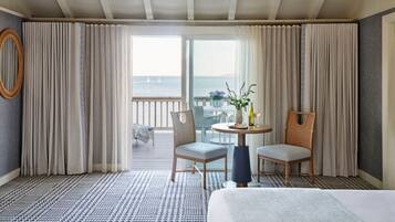 A hotel room with a large bed, two chairs, a small table with a vase and wine glasses, and a view of the sea through a sliding glass door.