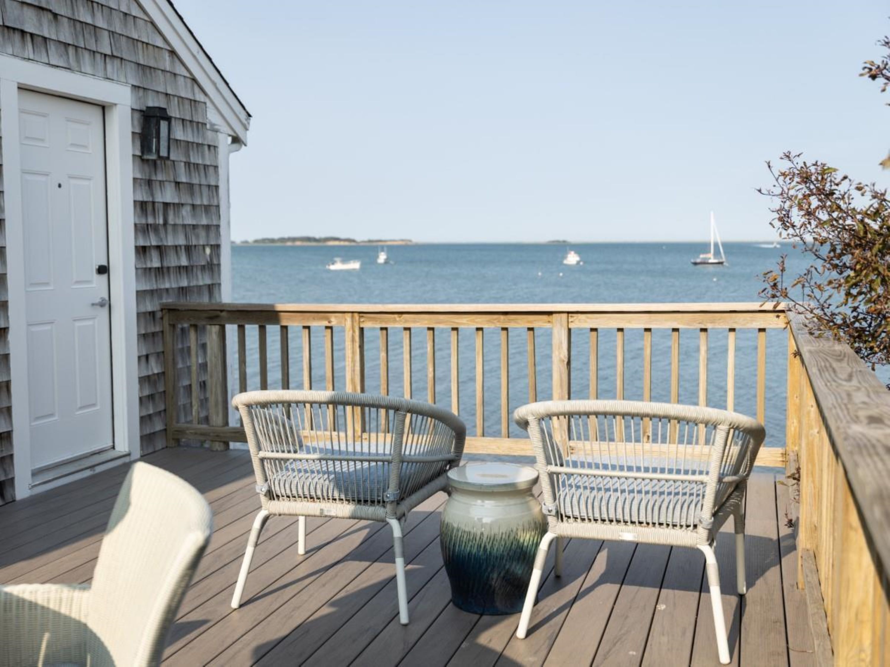 A wooden deck with two wicker chairs and a table, overlooking a body of water with boats.