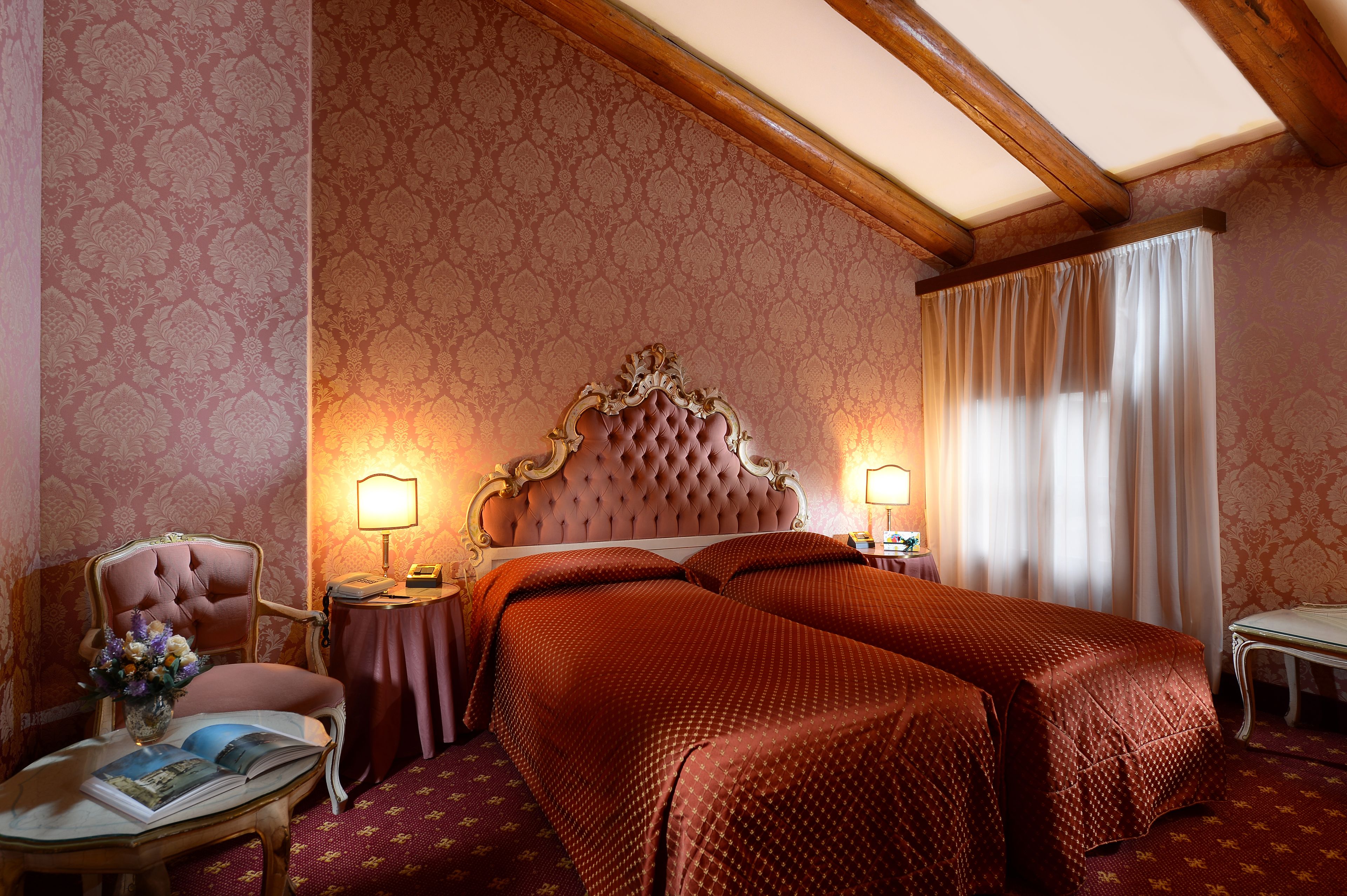 Habitación de hotel con una cama grande, dos mesitas de noche con lámparas, una silla y una pequeña mesa con un libro y flores.