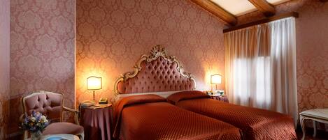 Habitación de hotel con una cama grande, dos mesitas de noche con lámparas, una silla y una pequeña mesa con un libro y flores.