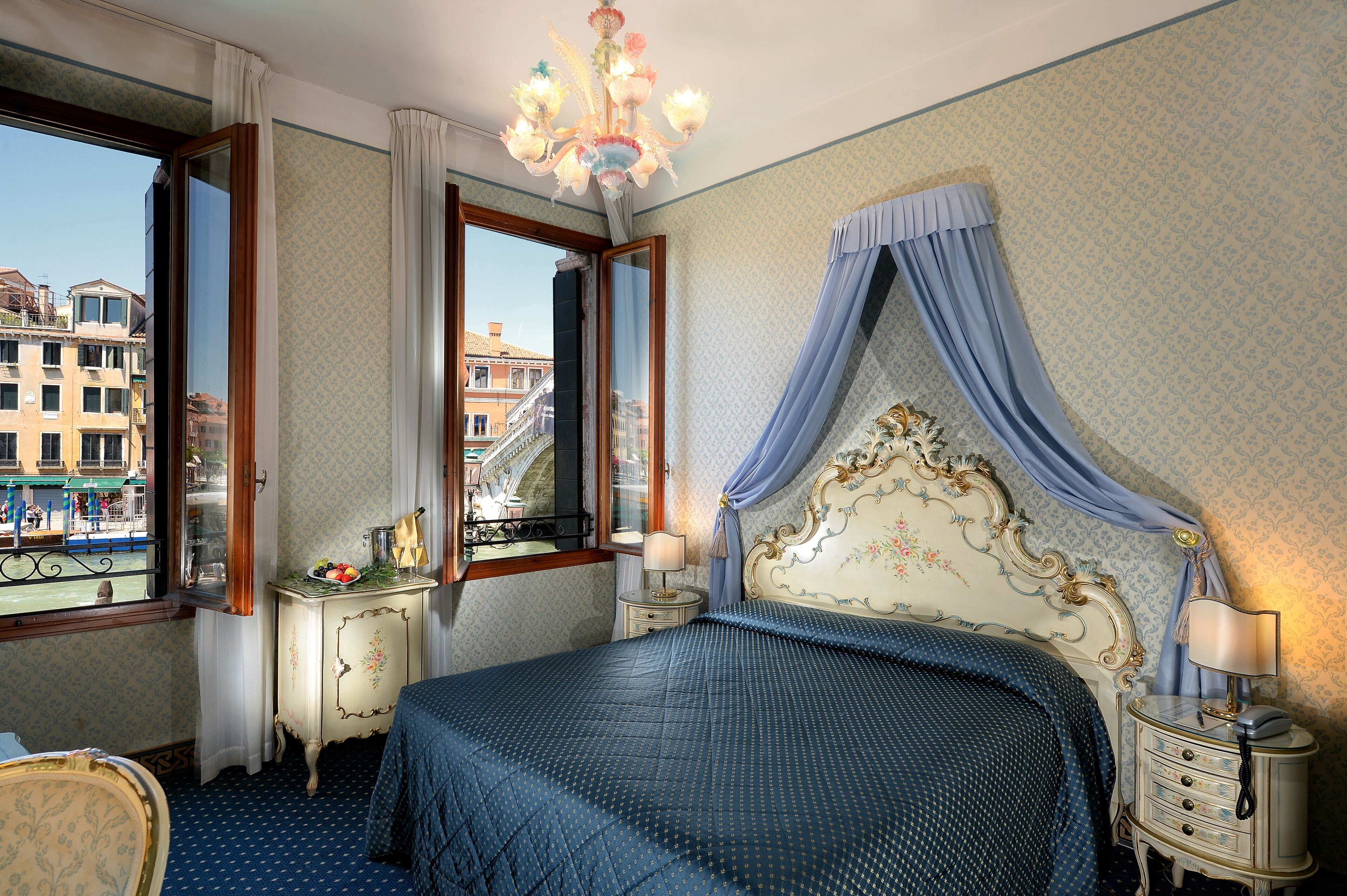 Un dormitorio con una cama grande, una mesita de noche, un aparador, una araña y una ventana con vistas a edificios.