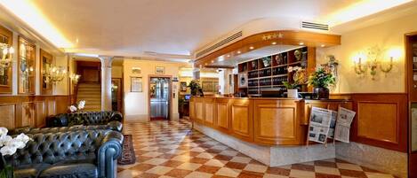A hotel lobby with a checkered floor, a leather sofa, a reception desk, and a staircase in the background.