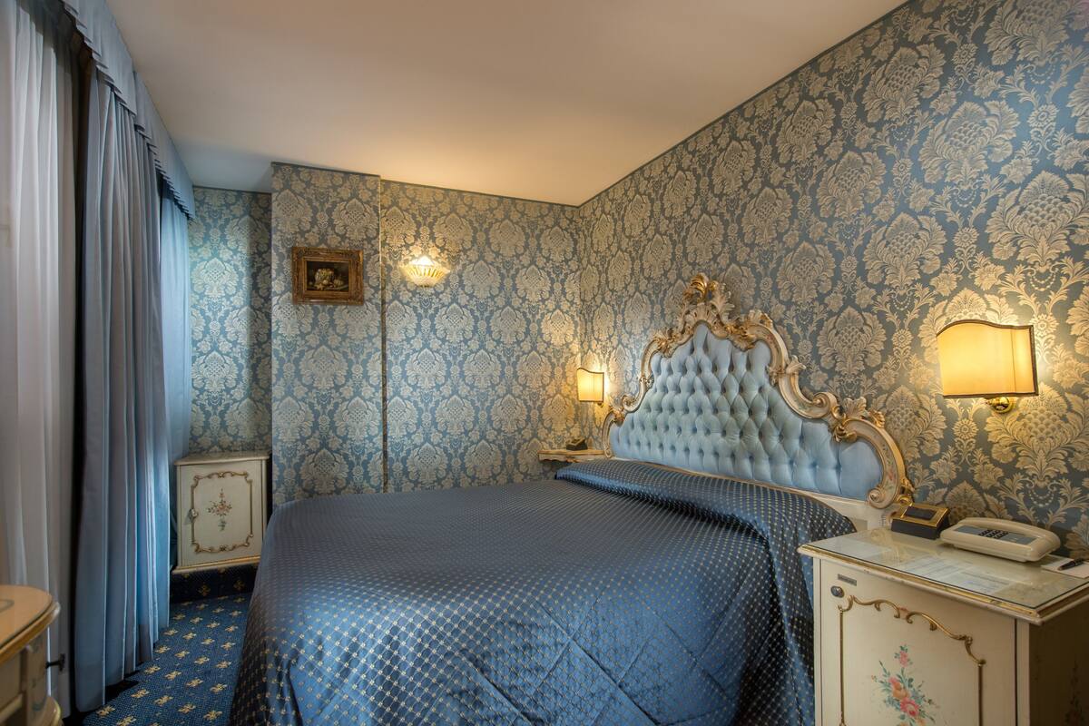 A bedroom with a large bed, ornate headboard, patterned wallpaper, and a nightstand with a lamp.