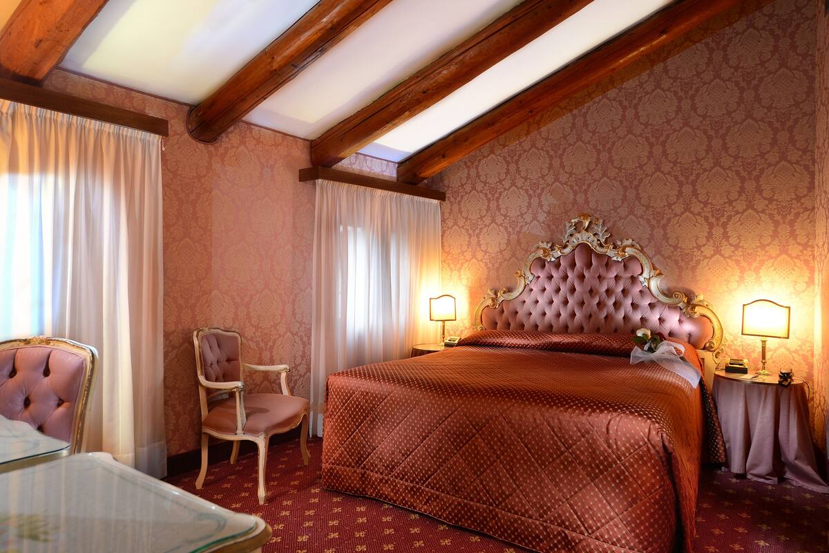 A room with a large bed, a chair, a table, and a nightstand with lamps.