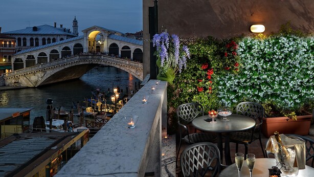 A terrace with a view of a historic bridge over a river, outdoor seating with tables and chairs, and a decorative wall with flowers. - Hotel Rialto (Venice)