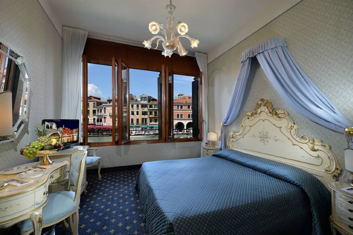 A hotel room with a bed, a desk with a chair, a television, and a view of buildings through the window.