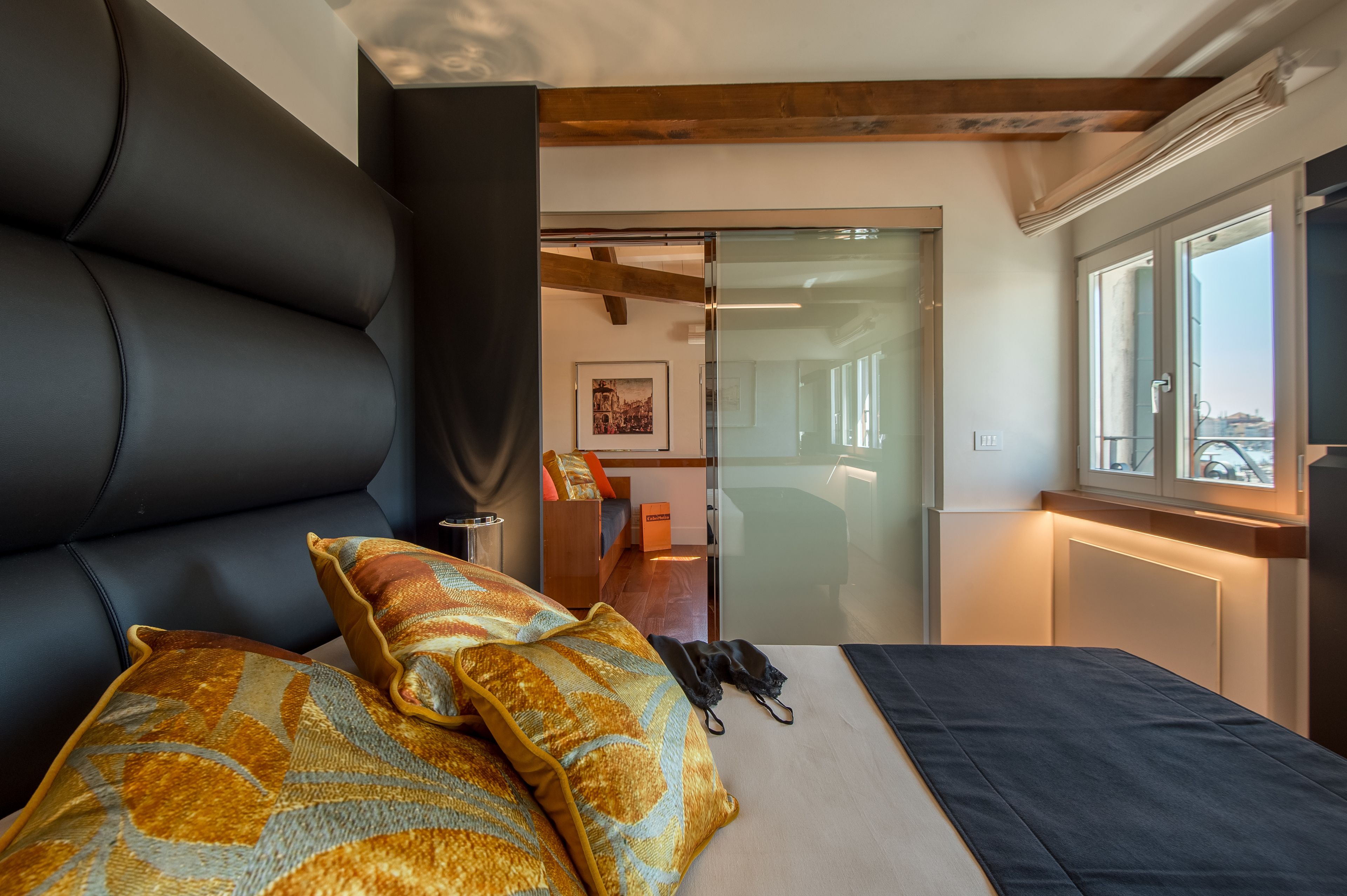 A modern bedroom with a large bed, two patterned pillows, a bedside table, and a view of the city through a window.
