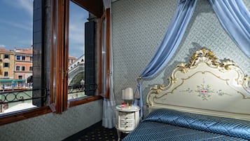 A room with a bed, a nightstand, a window with curtains, and a view of buildings outside.