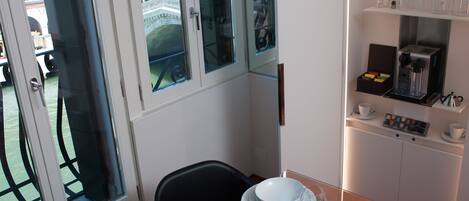 Un coin repas avec une table dressée pour deux personnes, une machine à café et une vue sur un immeuble à travers une porte vitrée.