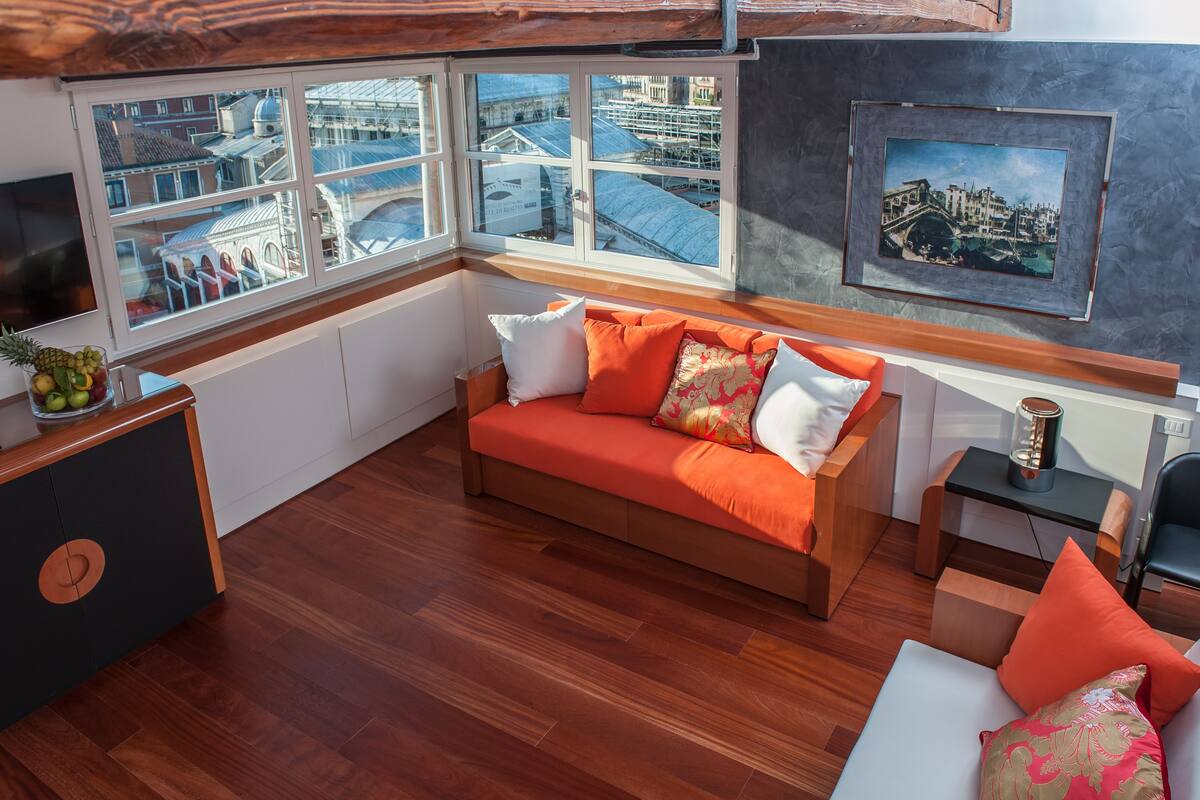 A modern room with a wooden floor, a sofa with orange cushions, a small table, and a framed picture on the wall.