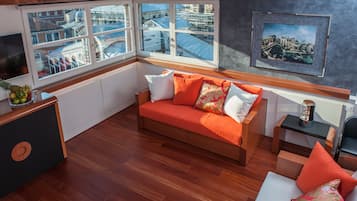 Una habitación moderna con suelo de madera, un sofá con cojines naranja, una mesita y un cuadro en la pared.