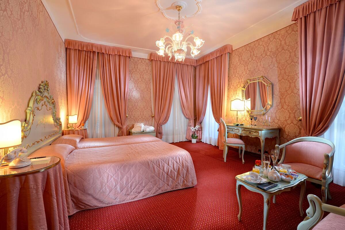 A hotel room with two beds, a vanity with a mirror, a dining table set for breakfast, and a chandelier.