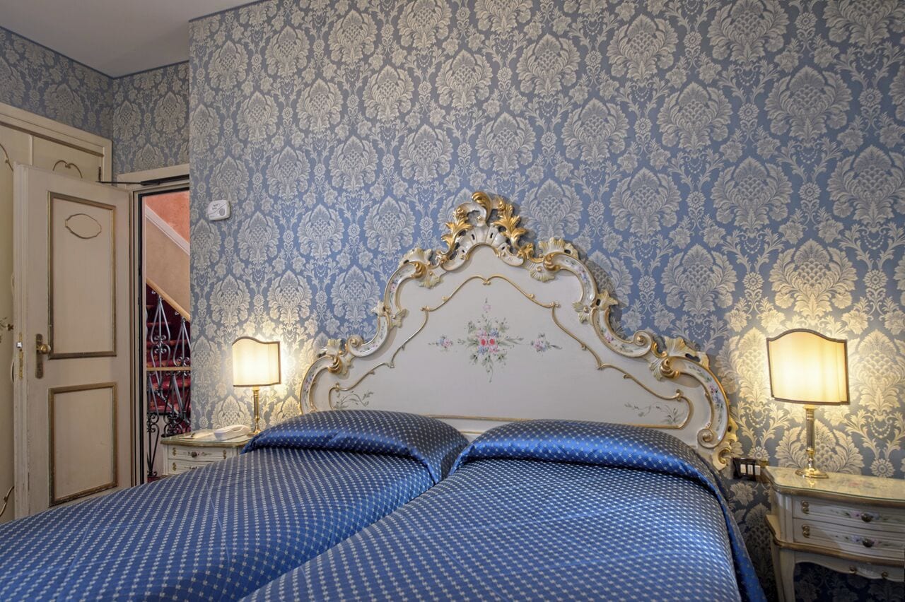 Un dormitorio con una cama grande y decorativa, dos mesitas de noche con lámparas, y un papel tapiz estampado.