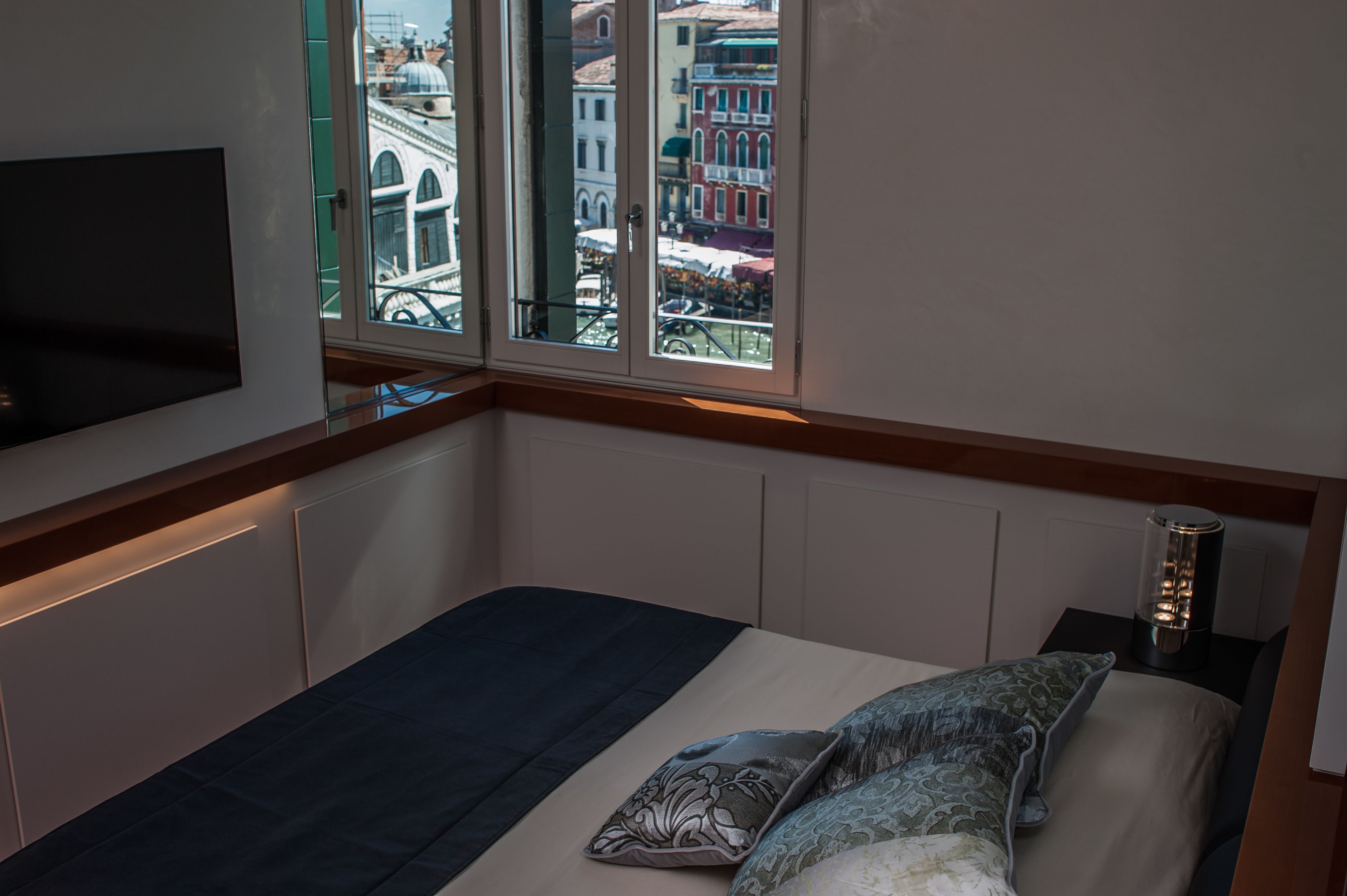 A bedroom with a bed, a TV mounted on the wall, and a view of a cityscape through a window.