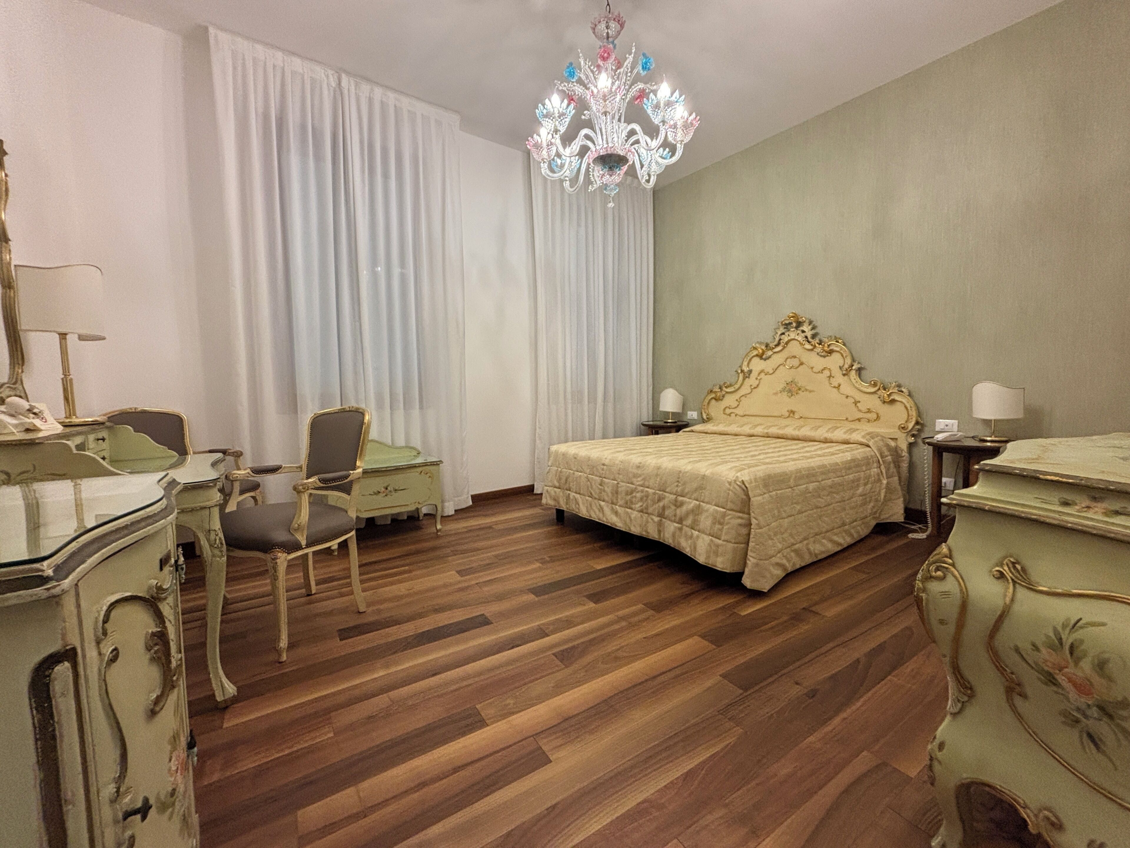 Una habitación de hotel con piso de madera, una cama con cabecera dorada, un escritorio con silla y un candelabro.