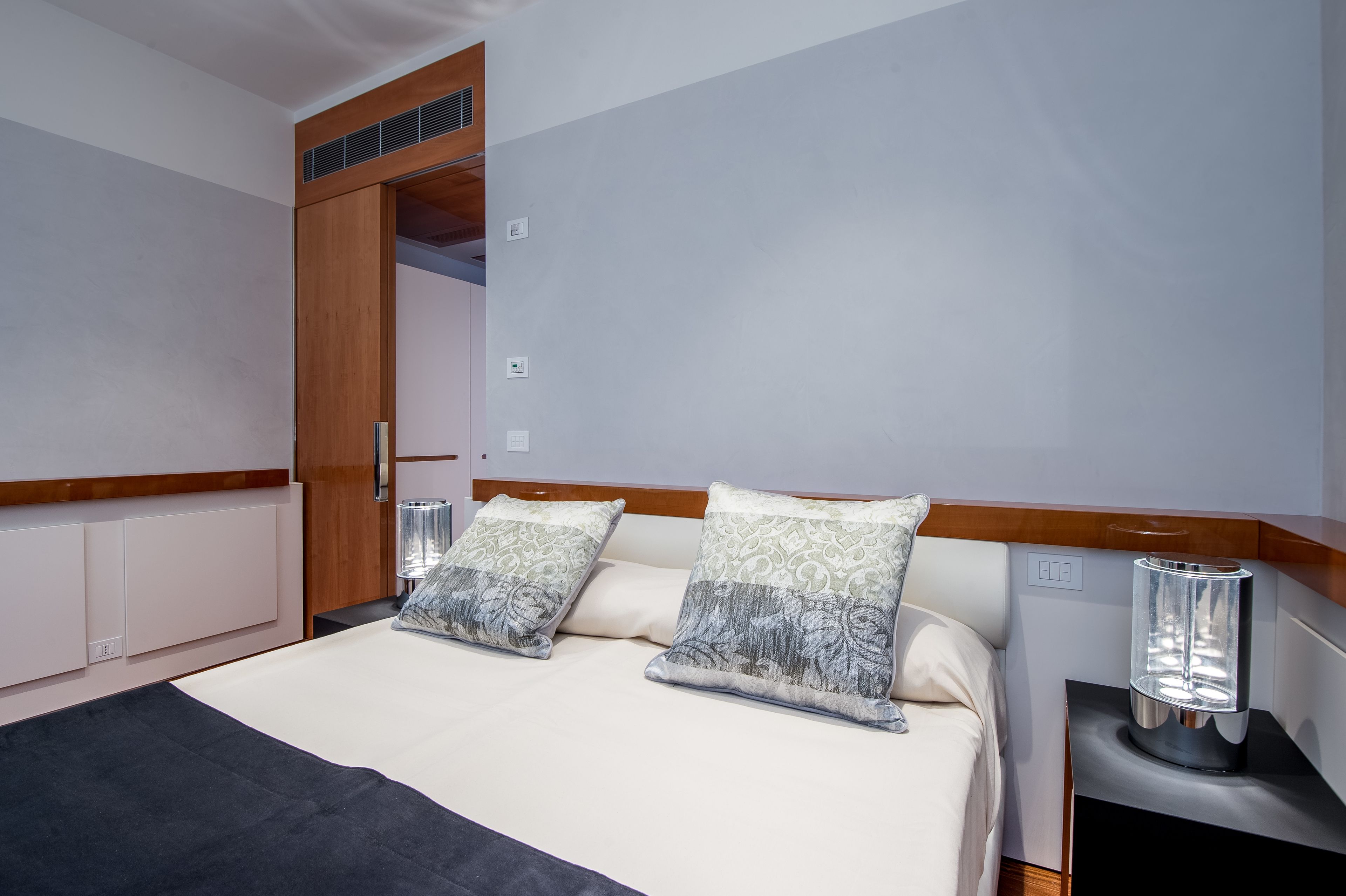A bedroom with a bed, two decorative pillows, a bedside table with a lamp, and a wardrobe.