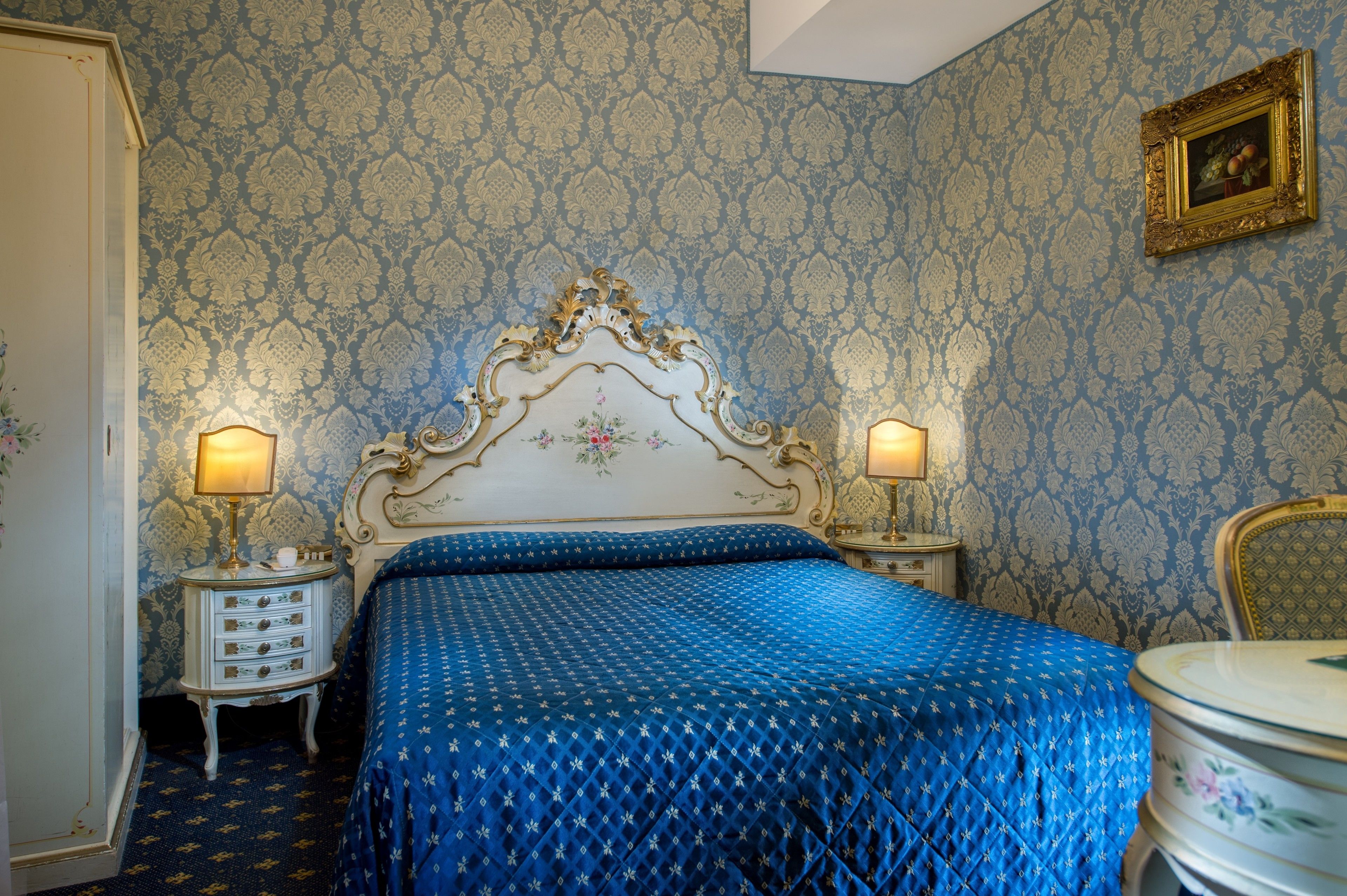 A room with a patterned wallpaper, a classic bed with a decorative headboard, two bedside tables with lamps, and a chair.