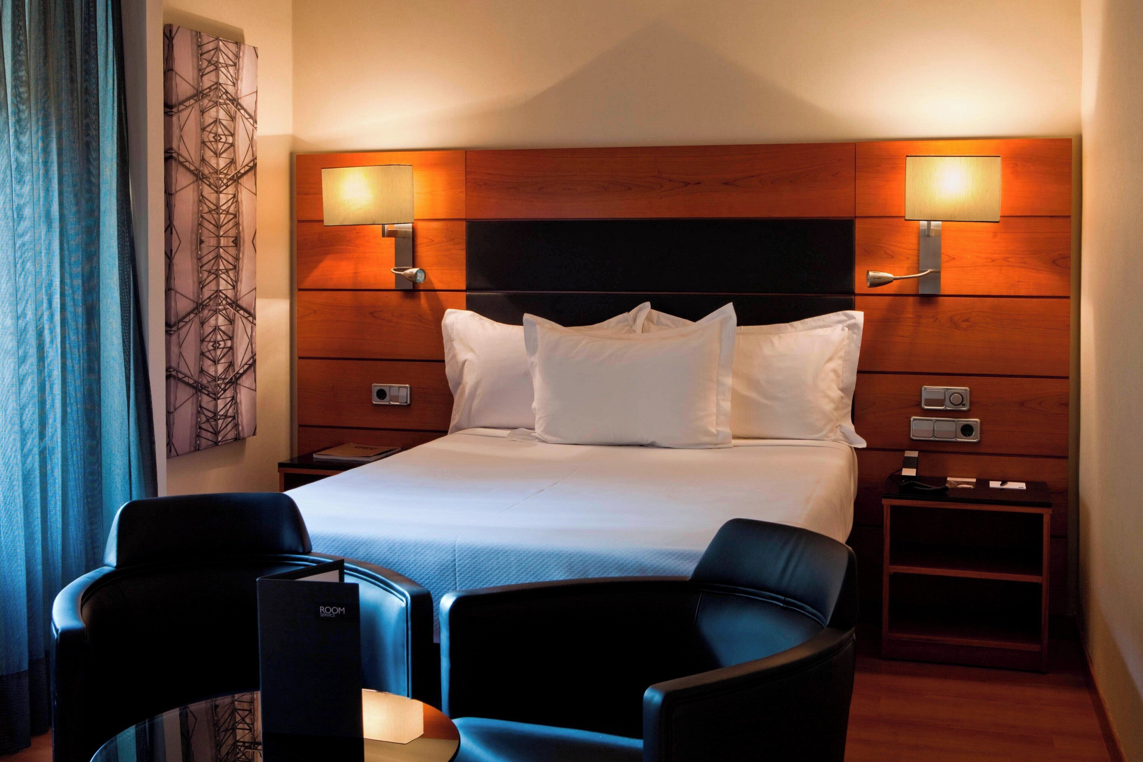 A hotel room with a large bed, two bedside tables with lamps, a chair, and a glass table.