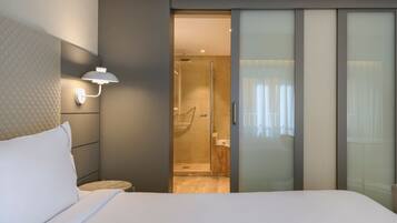 Una habitación de hotel moderna con una cama grande, una mesita de noche, una lámpara de pared y una puerta corrediza de vidrio que da a una zona de ducha.