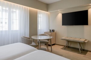 Ein modernes Hotelzimmer mit einem Bett, einem runden Tisch mit Stühlen, einer kleinen Kommode und einem flachbildfernseher an der Wand.