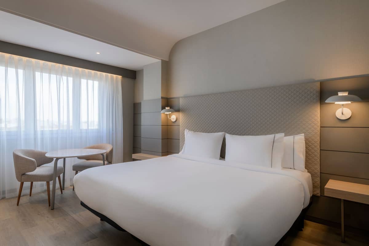 Ein Hotelzimmer mit einem großen Bett, zwei Stühlen, einem kleinen Tisch und einem Nachttisch mit einer Lampe.
