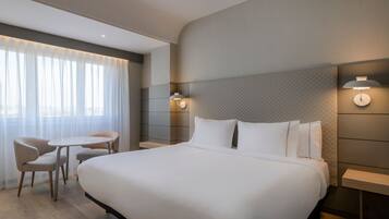 Una habitación de hotel con una cama grande, dos sillas, una mesita y una mesita de noche con lámpara.