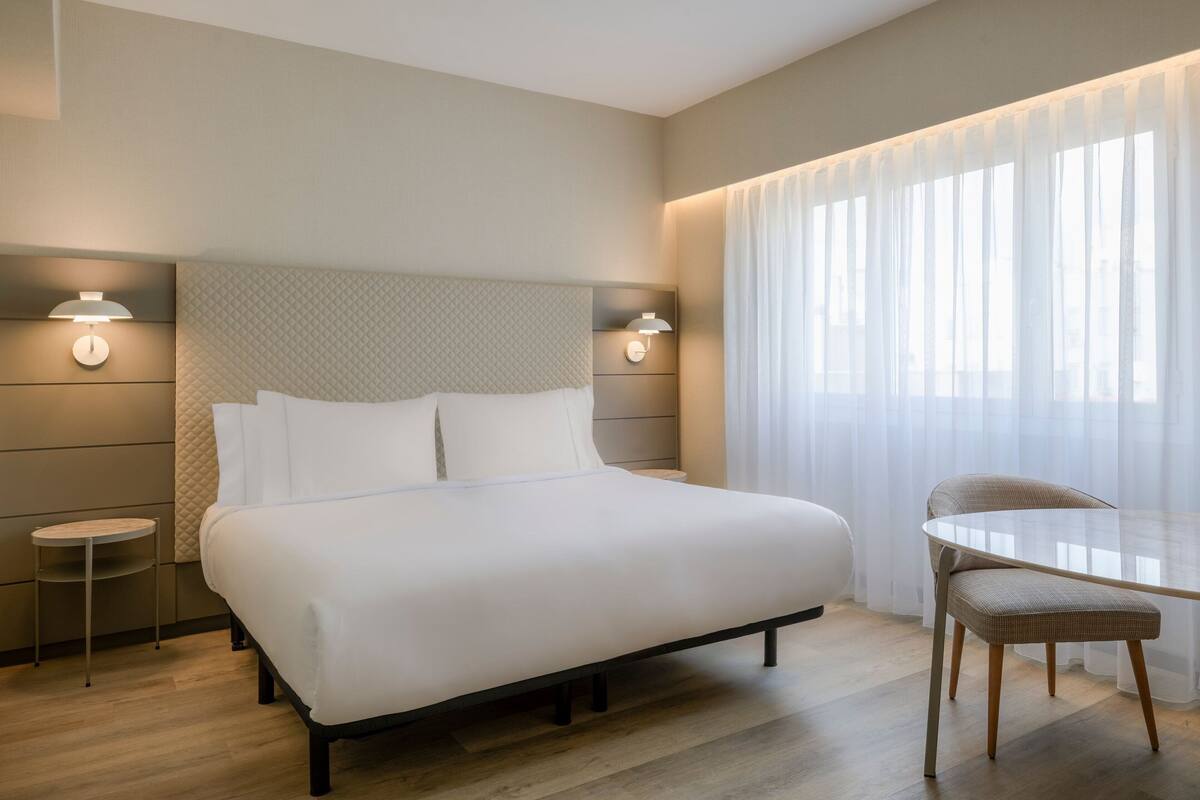 Ein modernes Hotelzimmer mit einem großen Bett, Nachttischen, einem kleinen runden Tisch und einem Sessel.