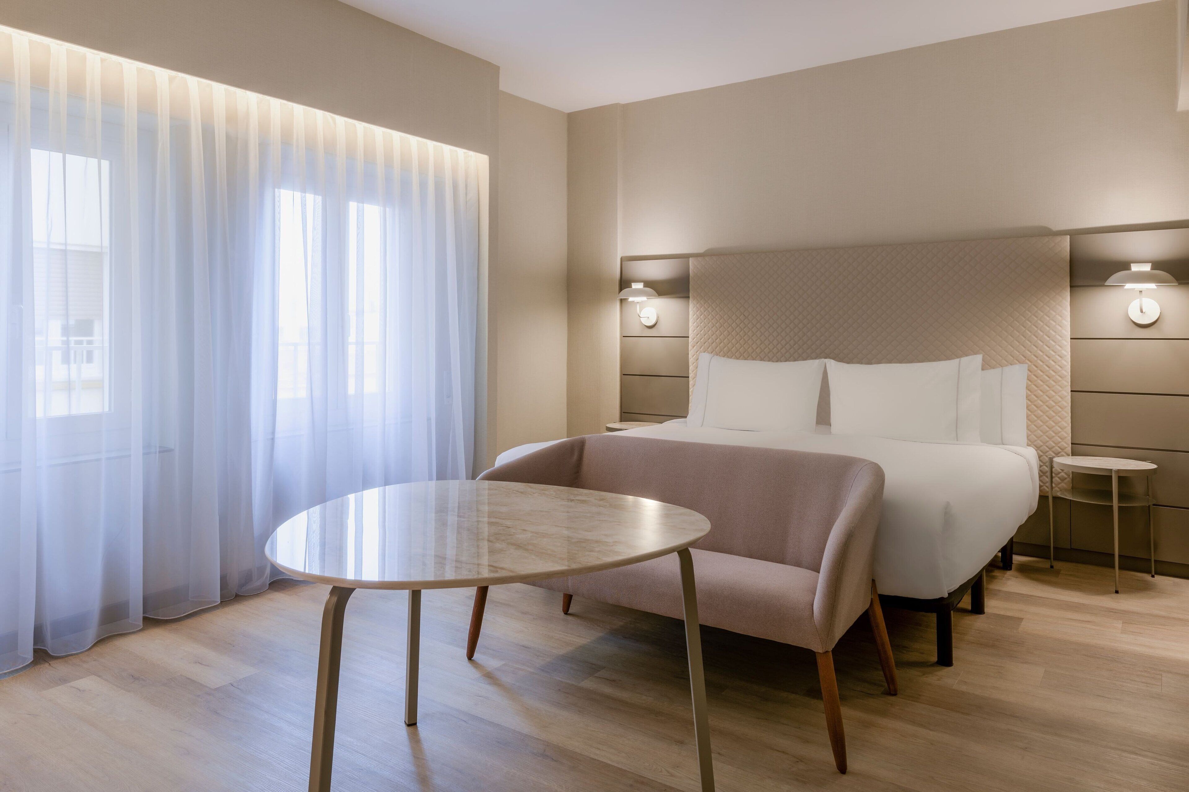 Een moderne hotelkamer met een groot bed, een ronde tafel en uitzicht op het landschap door dunne gordijnen.