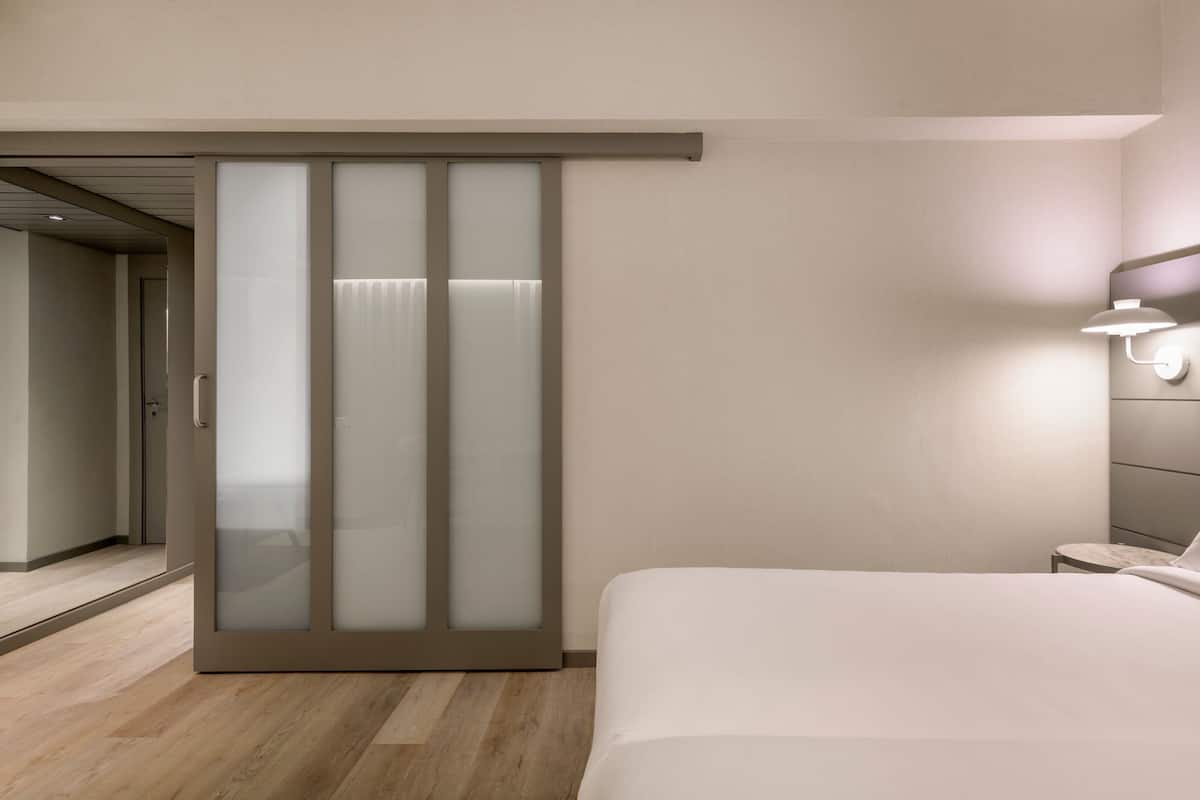Ein modernes Hotelzimmer mit einer Schiebetür, einem Bett, einem Nachttisch und einer wandmontierten Lampe.