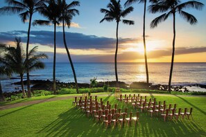 Montaje para una ceremonia de boda con sillas dispuestas en forma de corazón sobre un césped, con vista al océano, palmeras y un atardecer de fondo.