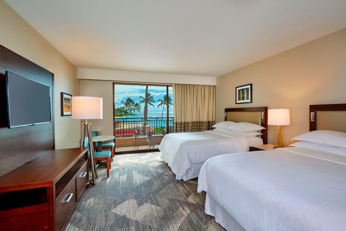 A hotel room with two beds, a desk, a chair, a TV, and a balcony with a view of palm trees and the ocean.