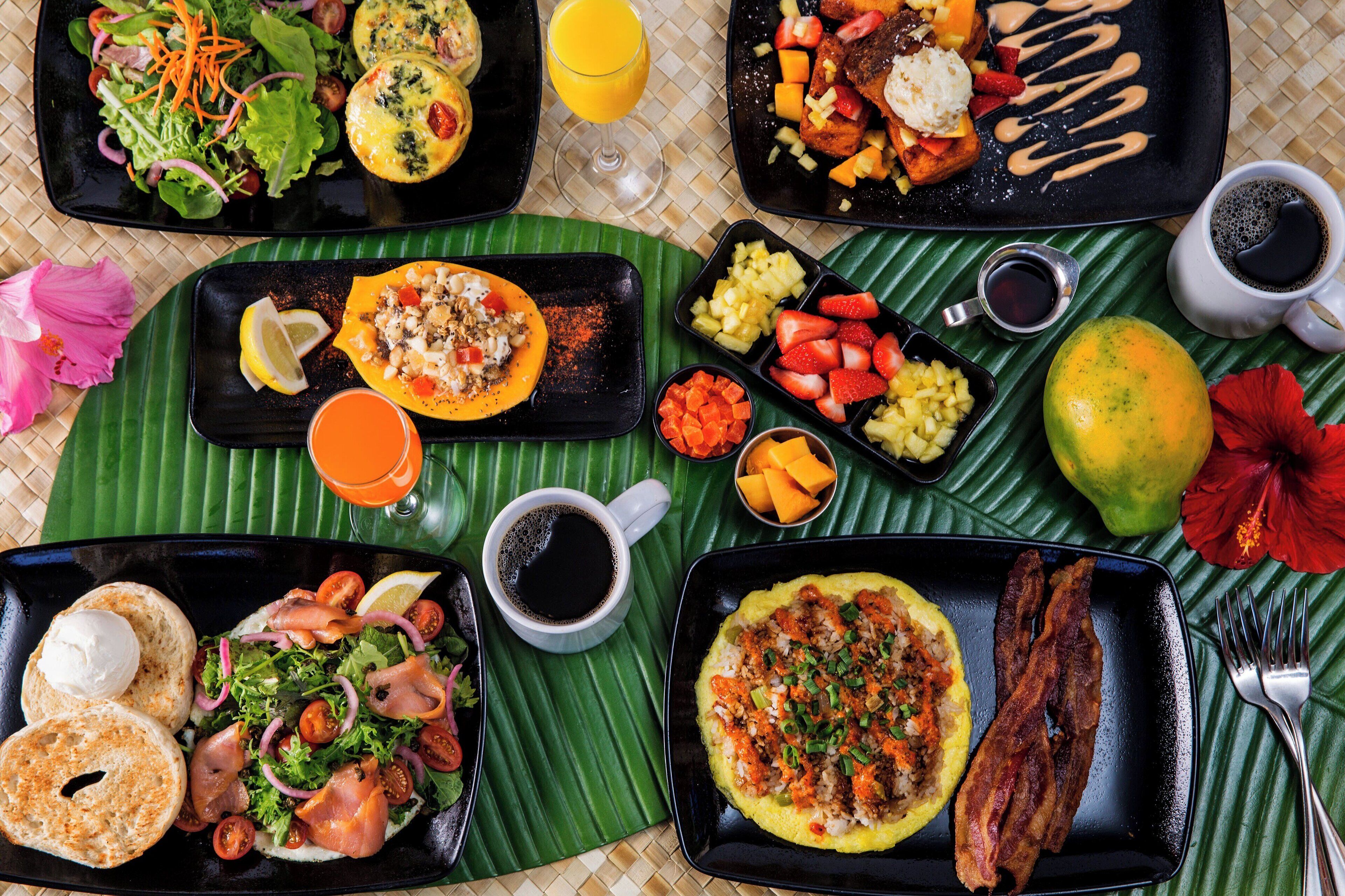 A variety of breakfast dishes on black plates, including eggs, bacon, and fresh fruits, arranged on a green mat.
