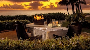 An outdoor dining setup with a table set for two, surrounded by chairs, a fire pit, and candles. - Sheraton Kauai Resort (Koloa)
