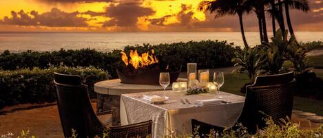 An outdoor dining setup with a table set for two, surrounded by chairs, a fire pit, and candles.