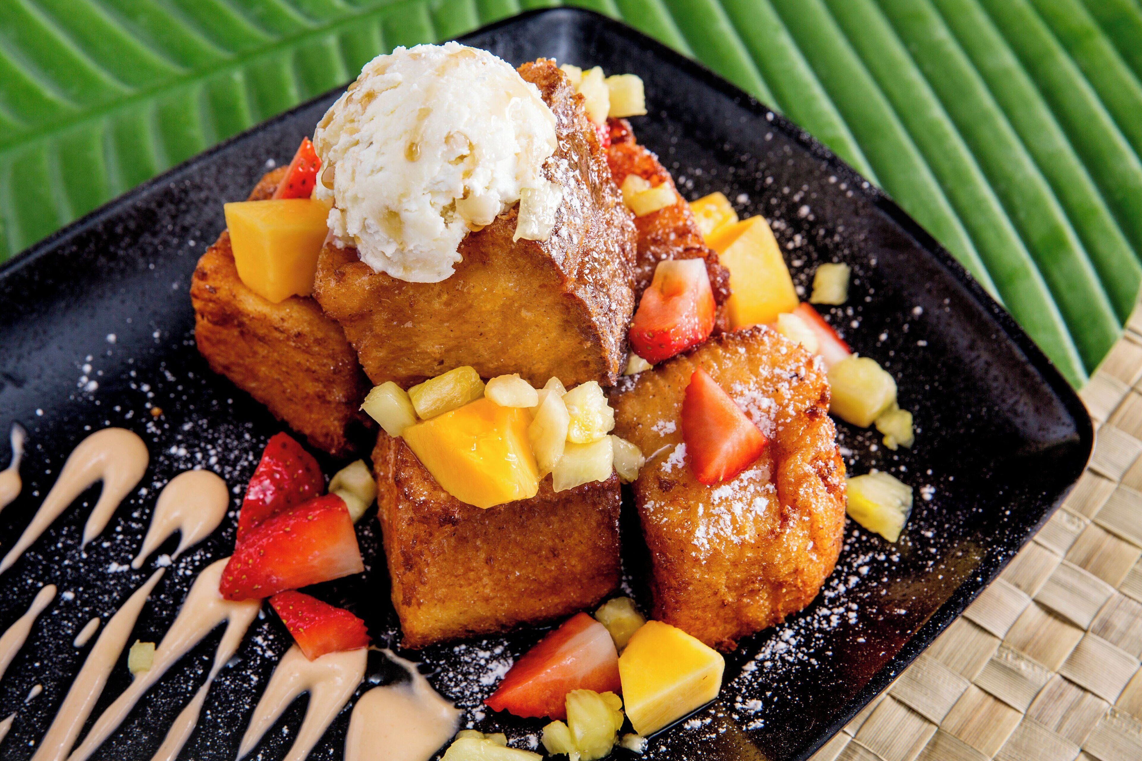 A square plate with a dessert consisting of a golden-brown item topped with ice cream and diced fruit, accompanied by a drizzle of sauce.