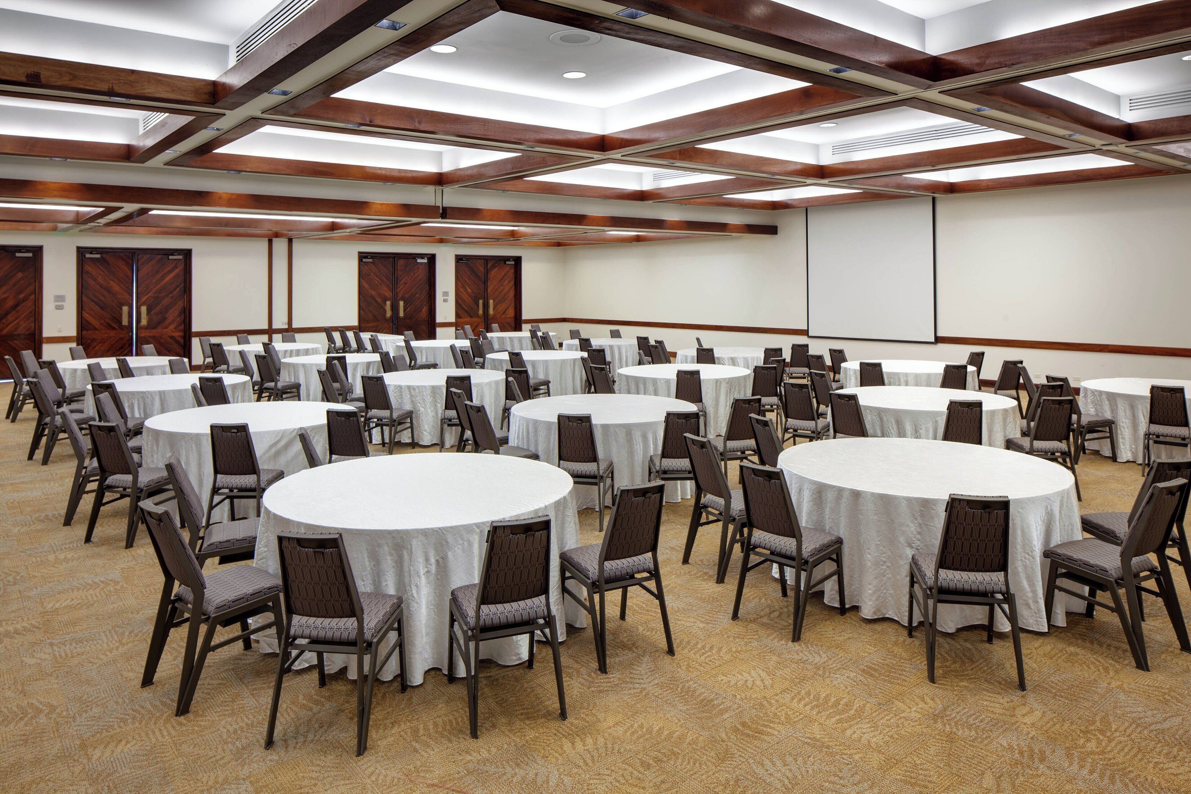 A conference room with multiple round tables and chairs, wooden paneling on the walls, and a large screen at the front.