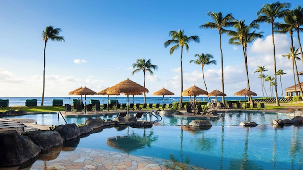 A pool area with lounge chairs and umbrellas, palm trees, and a view of the ocean. - Sheraton Kauai Resort (Koloa)