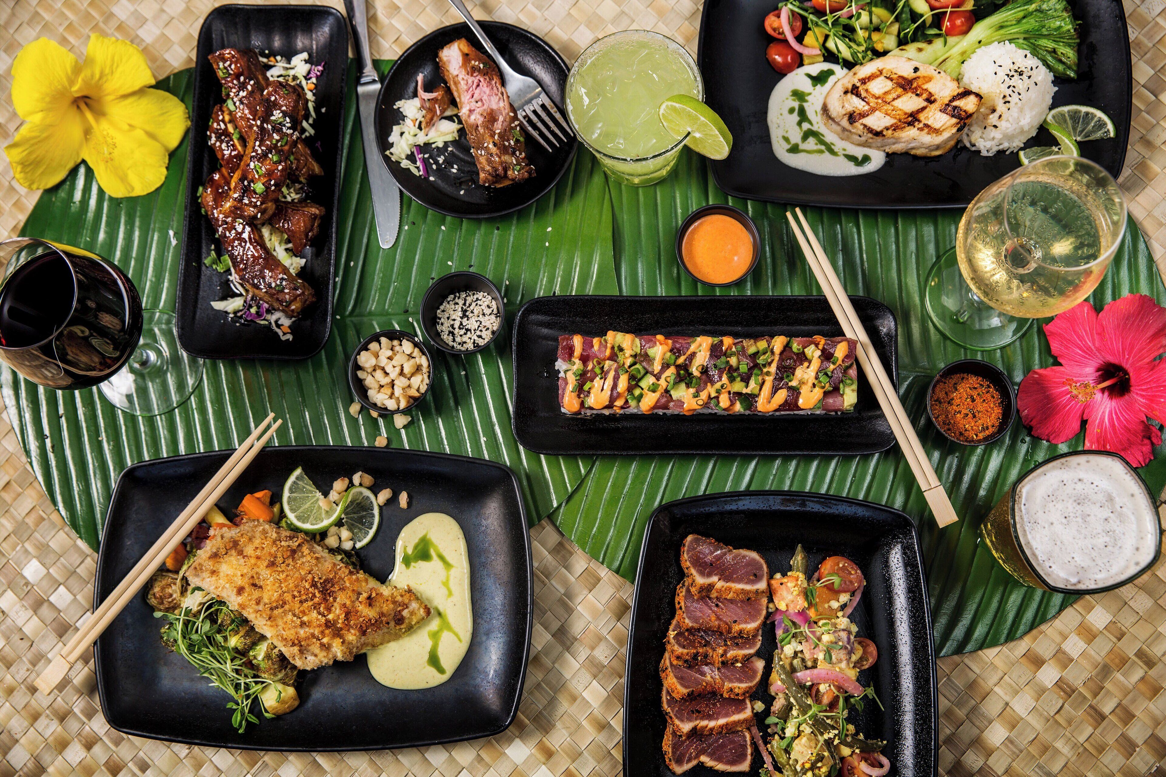 A variety of dishes including grilled meat, fish, and vegetables, served on black plates with a tropical flower and a drink.