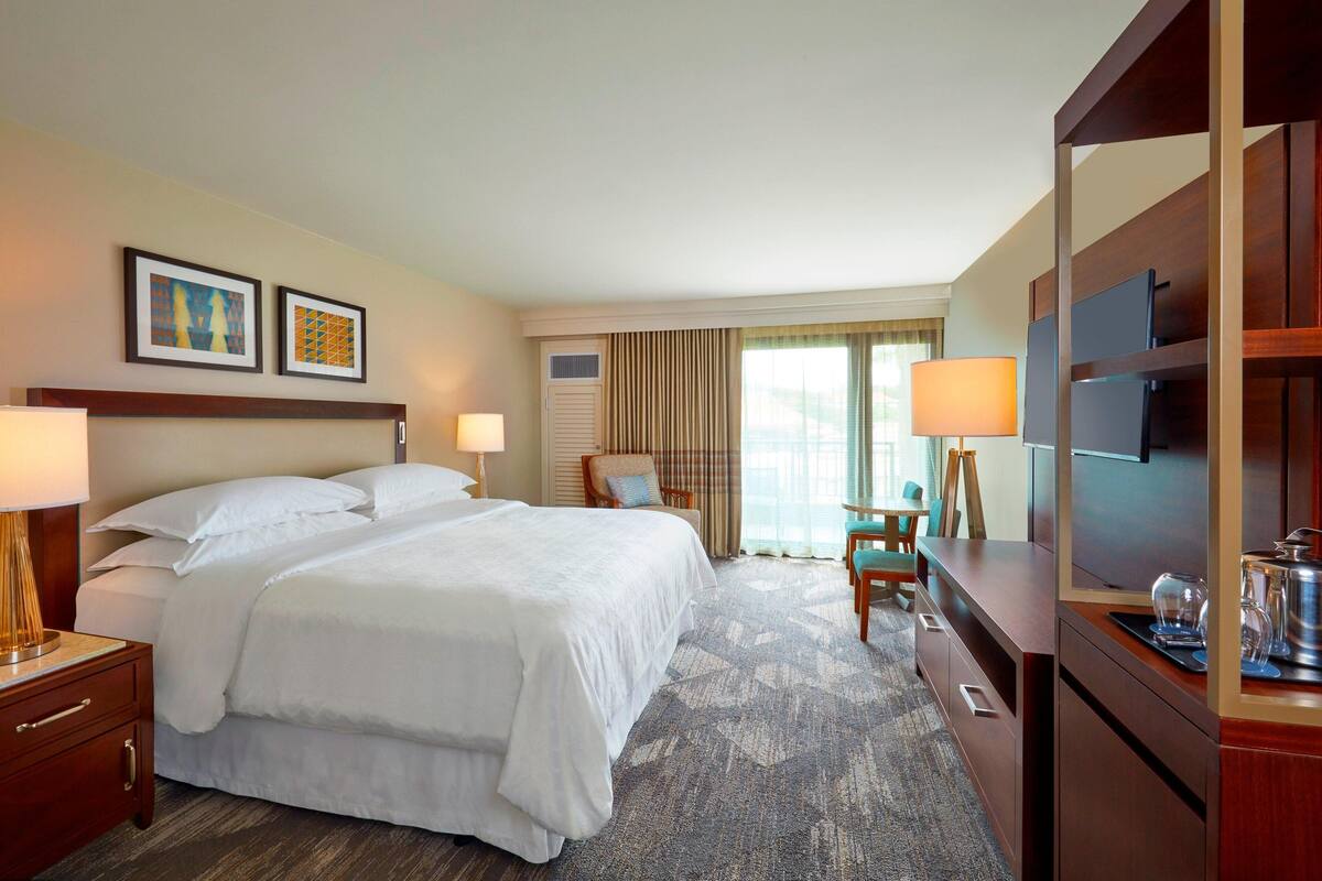 A hotel room with a large bed, a desk with a chair, a TV, and two framed pictures on the wall.