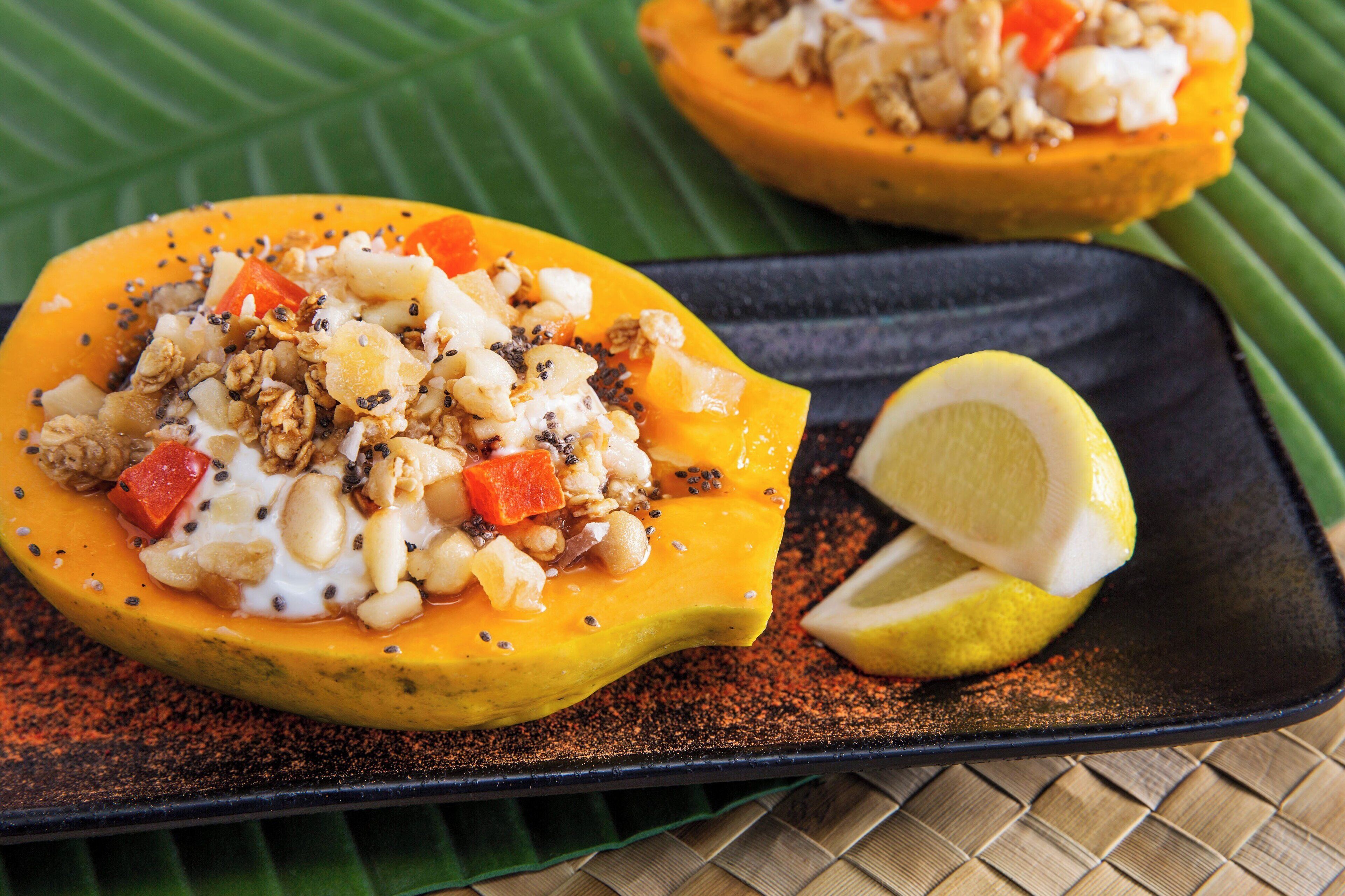 A dish with a halved papaya filled with a mixture of nuts, seeds, and possibly cheese, served on a black plate with a slice of lemon on the side.