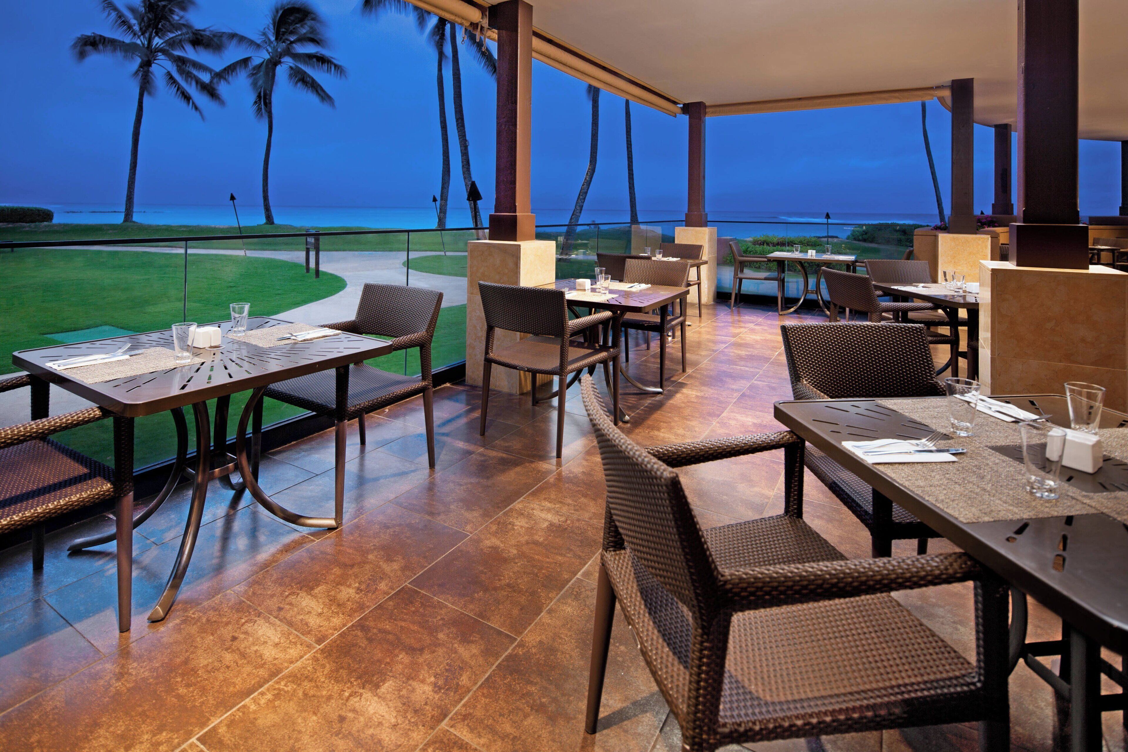 Un área de comedor al aire libre con mesas y sillas, con vista a un campo de golf y al océano al atardecer.