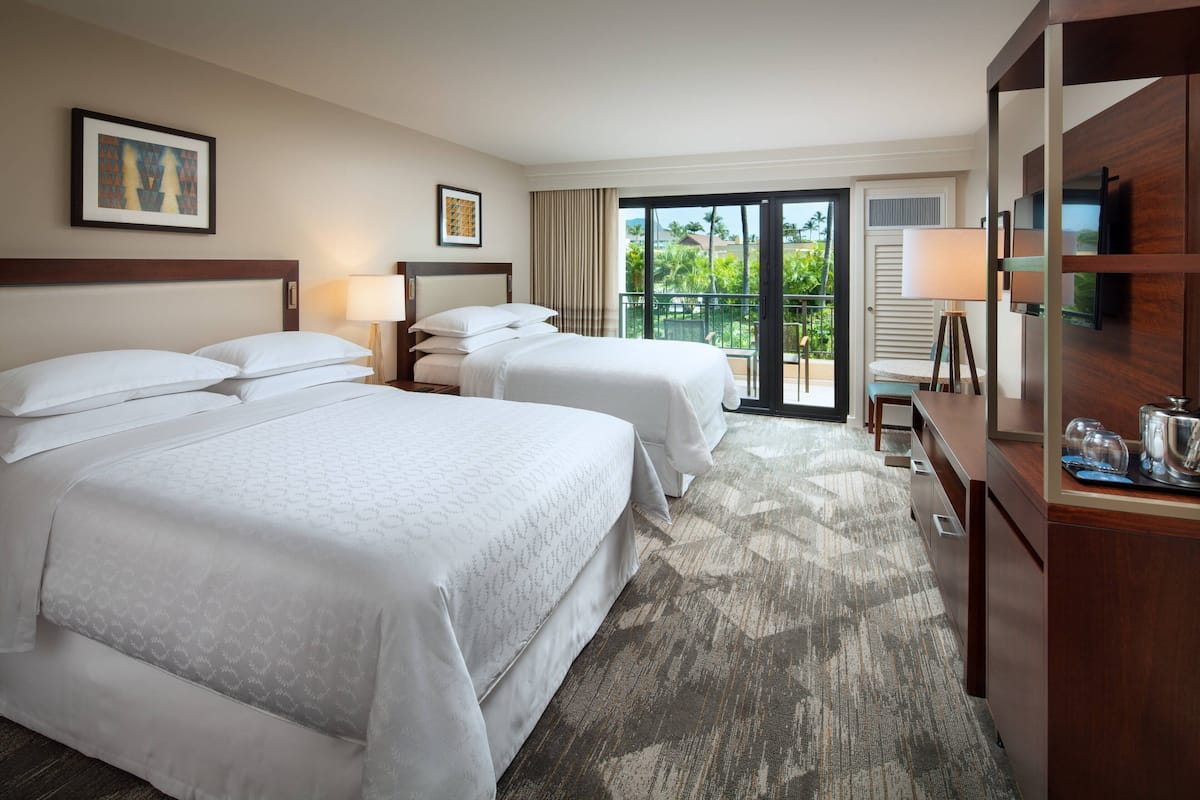 A hotel room with two beds, a desk, a TV, and a balcony with a view of palm trees.