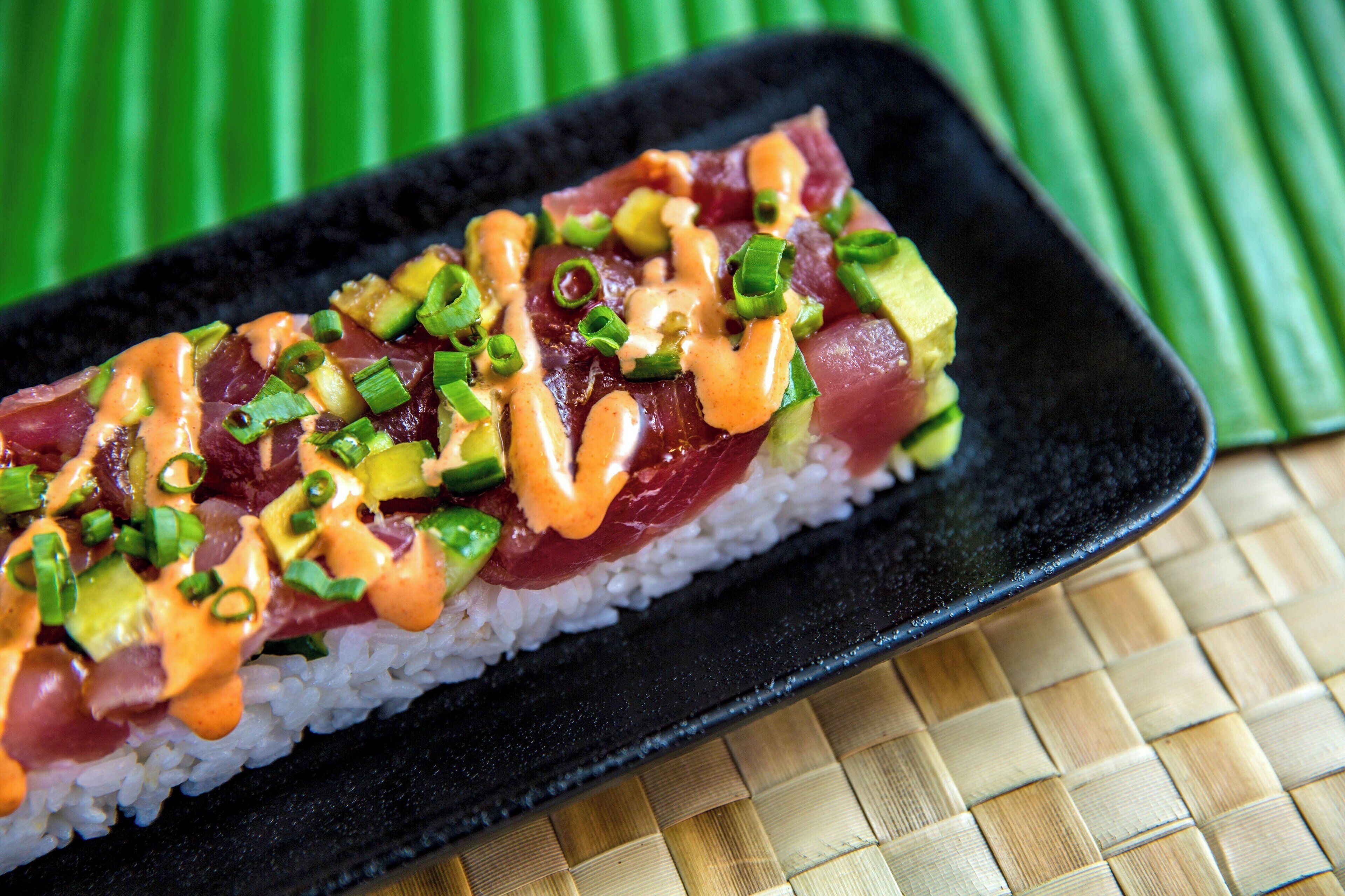 A sushi dish on a black plate with white rice, topped with avocado, green onions, and a sauce.