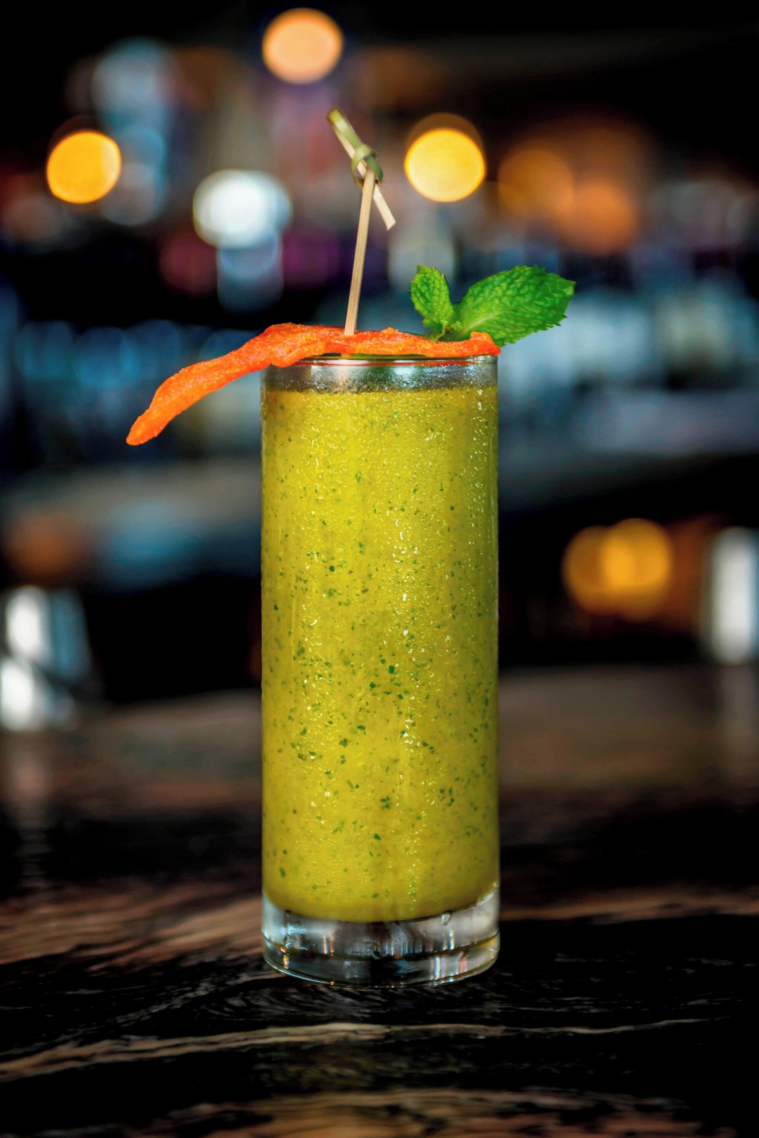 Un grand verre contenant une boisson verte, décoré d’une feuille de menthe et d’une tranche de carotte, posé sur le comptoir d’un bar.