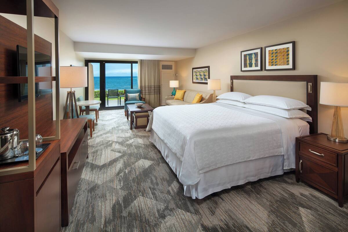 A hotel room with a large bed, a desk with a lamp, a chair, and a view of the ocean.