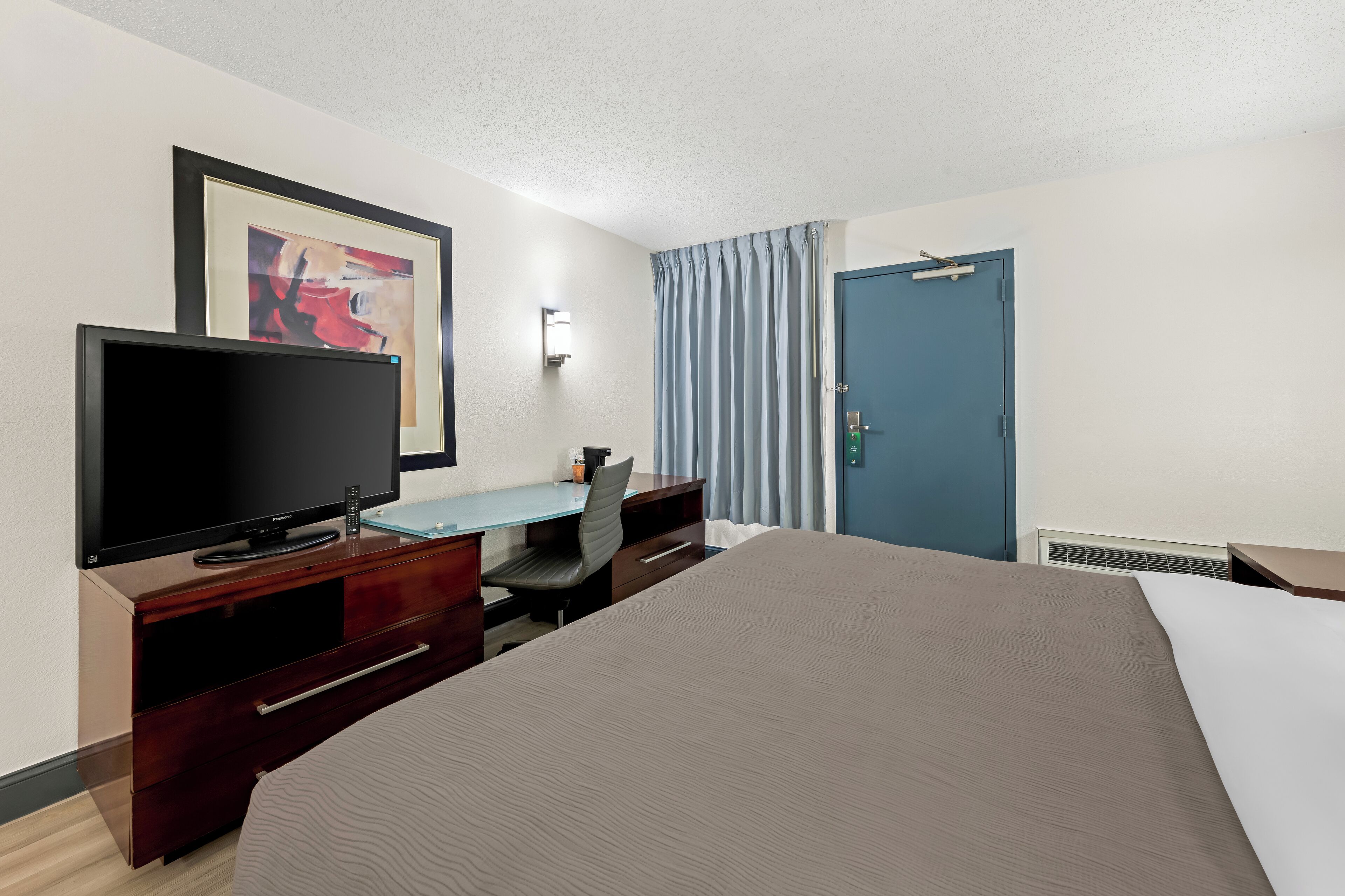 A hotel room with a bed, a desk with a chair, a television, a framed artwork, a door, and a window with curtains.