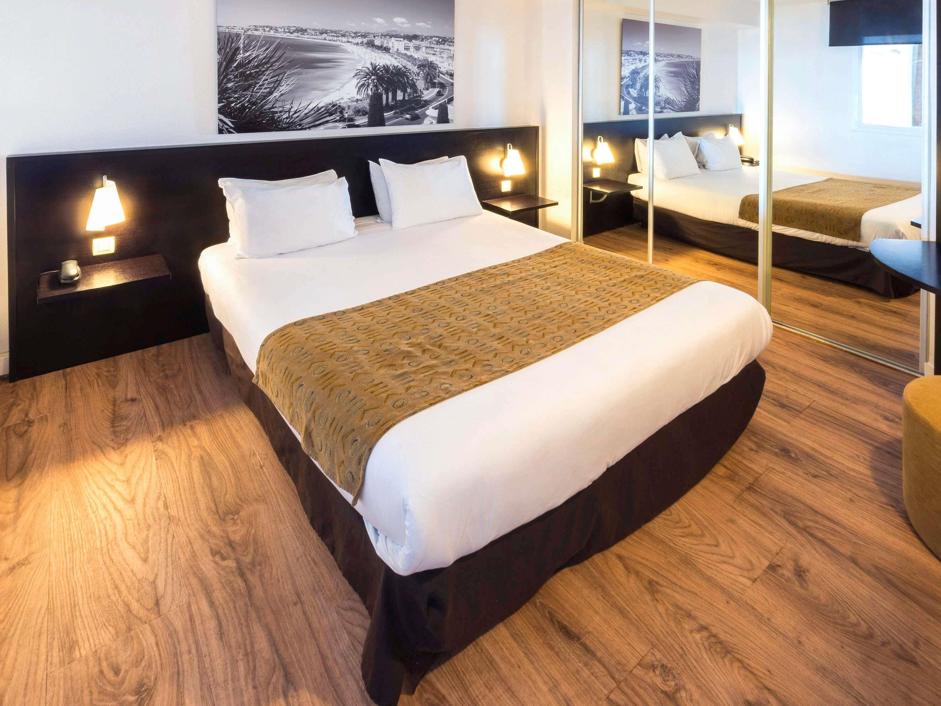 A hotel room with a large bed, bedside tables with lamps, a mirror, and a cityscape view.