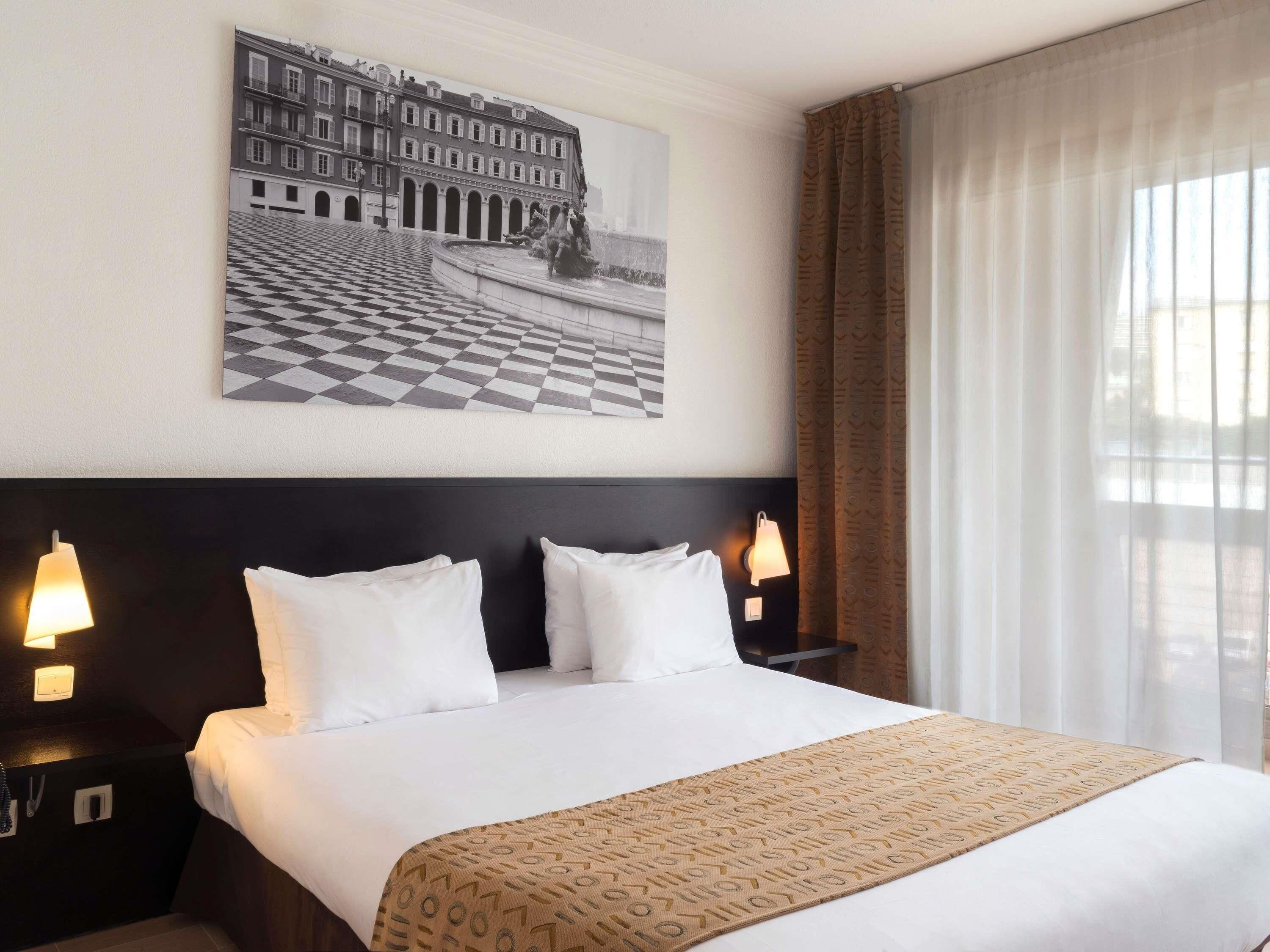Ein Hotelzimmer mit einem großen Bett, zwei Nachttischen mit Lampen, einem Schachbrettfußboden, einer gemusterten Tagesdecke und einem Stadtbild über dem Bett.