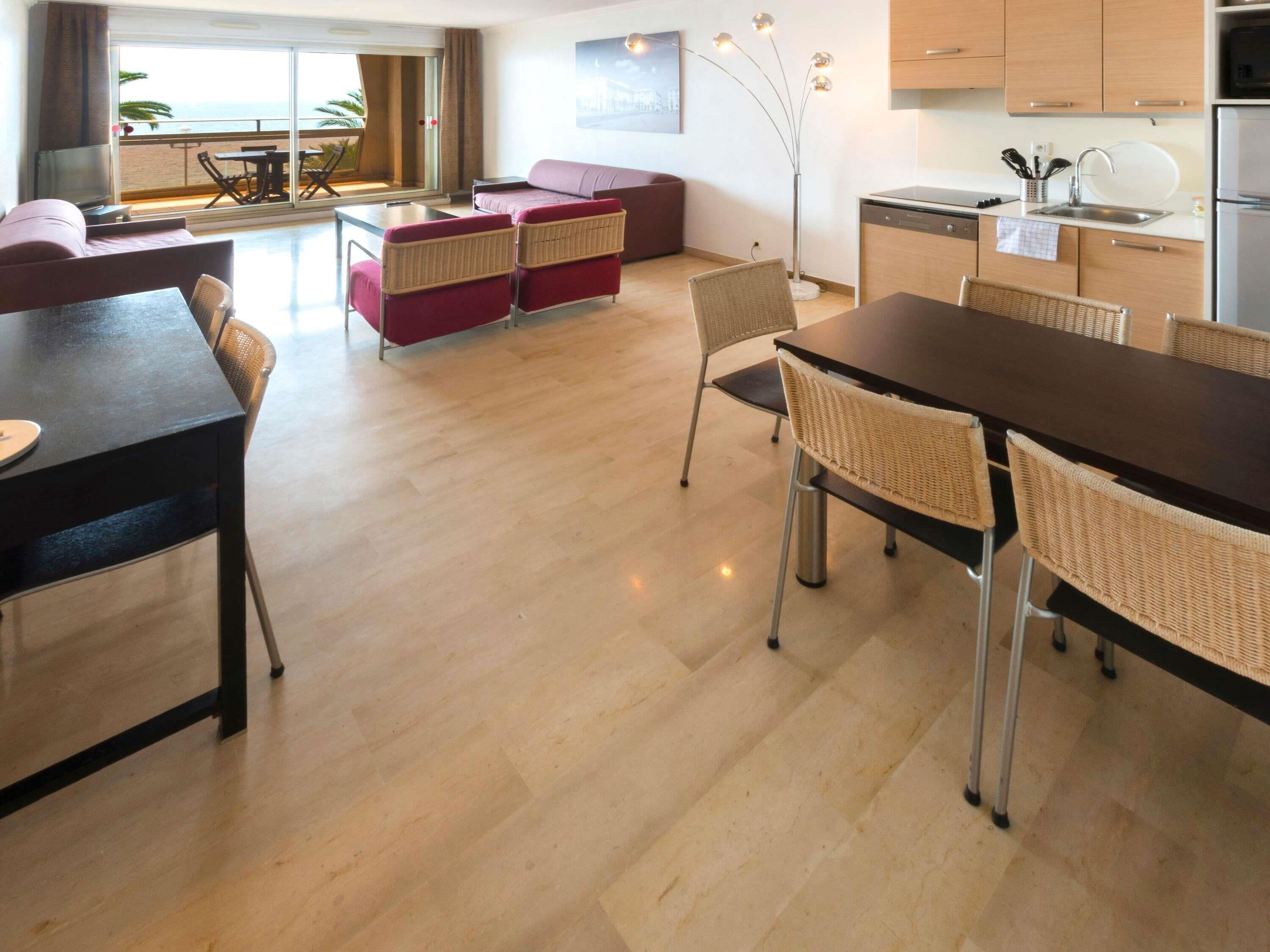 A modern hotel room with a dining area, a sofa, a kitchen, and a view of the sea.